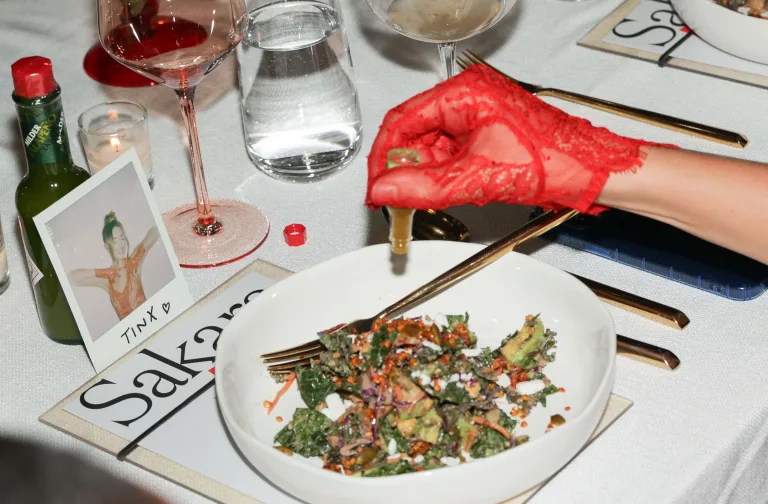 Overhead shot of a dining table with a salad being topped with Tabasco sauce by a hand wearing a red lace glove.