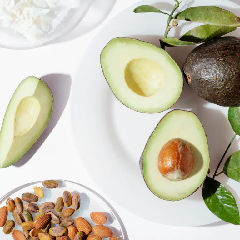 Halved avocados with pits, whole avocado, almonds, pistachios, and leafy greens arranged on a white plate.