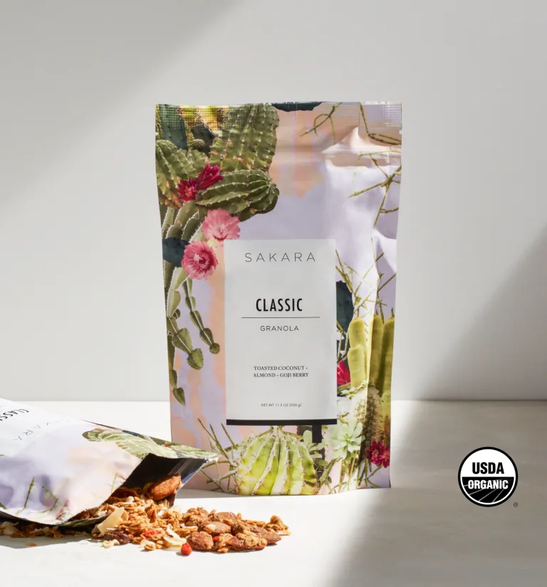 Sakara Classic Superfood Granola in resealable pouch with cactus and floral print, toasted coconut almond goji berry flavor, 11.5 oz (326 g), USDA Organic certified, shown with granola spilling onto surface.