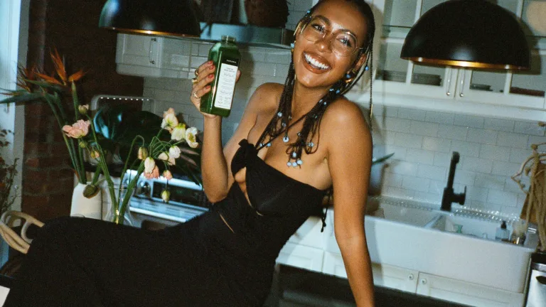 Hannah Traore sitting in a modern kitchen holding a green juice bottle and smiling.