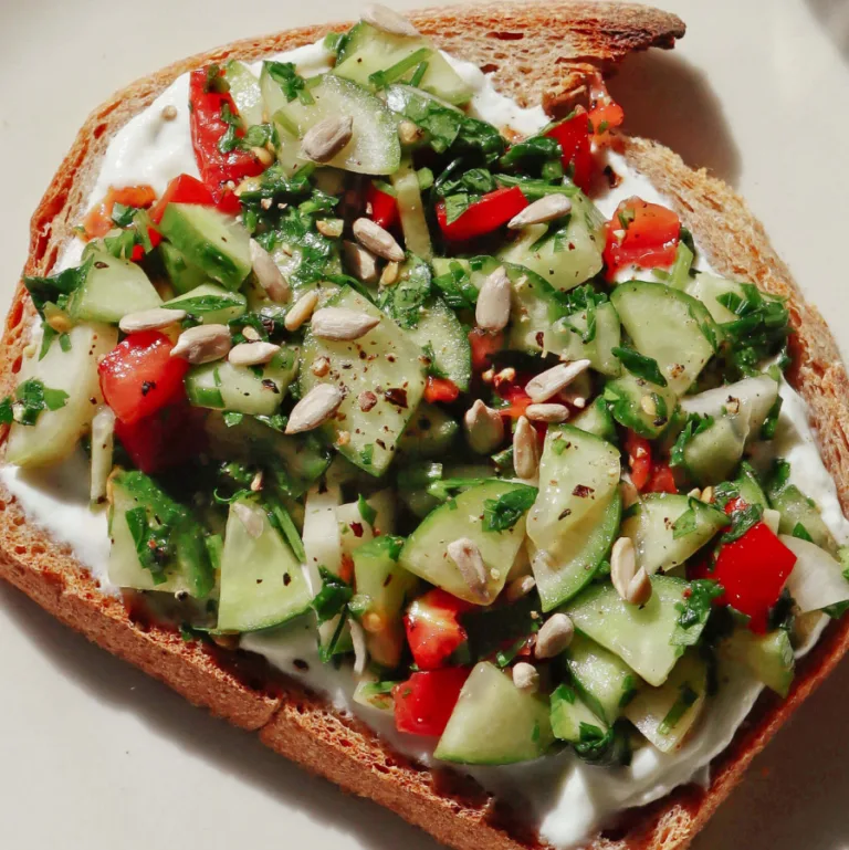 Close-up of mediterranean toast topped with chopped cucumber, red pepper, herbs, and sunflower seeds.