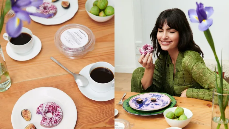 Woman in a green blouse sitting at a wooden table smiling while holding a pink-frosted Sakara breakfast donut, with coffee cups and small bowls on the table.