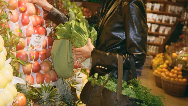 Person shopping in a grocery store holding leafy greens beside a display of tomatoes and fresh produce.