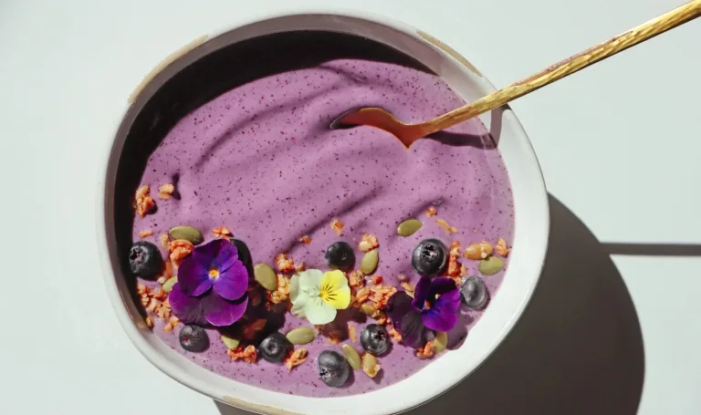 Purple smoothie bowl topped with blueberries, seeds, granola, and edible flowers with a gold spoon.