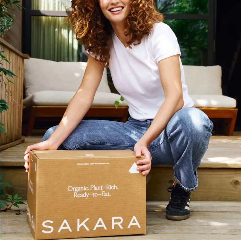 Sakara Signature Nutrition Program brown insulated delivery box labeled “Organic. Plant-Rich. Ready-to-Eat.” and “Sakara” held outdoors by a person.