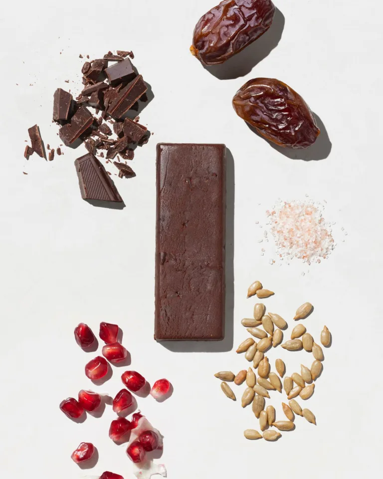 Sakara Metabolism Protein Super Bar in chocolate sea salt flavor shown unwrapped with chocolate, dates, sea salt, sunflower seeds, and pomegranate ingredients displayed around the bar.