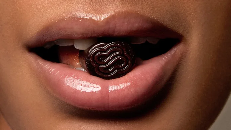 Close-up glossy lips holding a dark purple gummy supplement between teeth (Sakara Metabolism Bite).