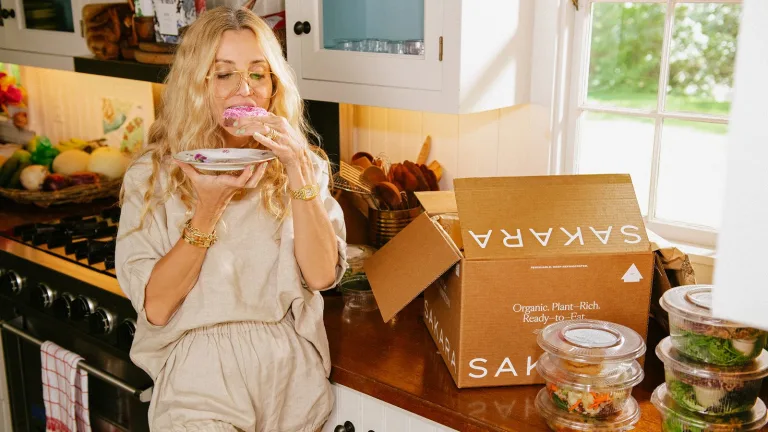 Woman standing in a kitchen eating a donut beside a brown delivery box labeled Sakara and stacked meal containers.
