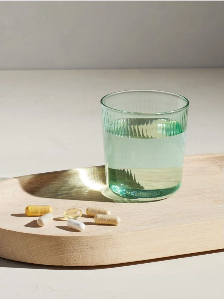 Sakara The Foundation assorted capsules and softgels displayed beside a green glass of water on a wooden tray with soft natural light.