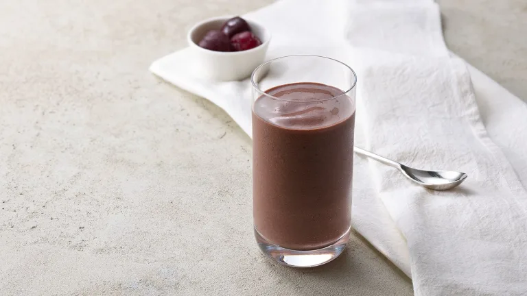 Glass of chocolate almond butter protein smoothie on a neutral surface with a white napkin and cherries in the background.