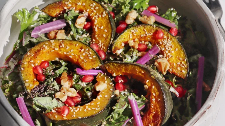 Salad topped with roasted squash slices, pomegranate seeds, and greens in a shallow bowl.